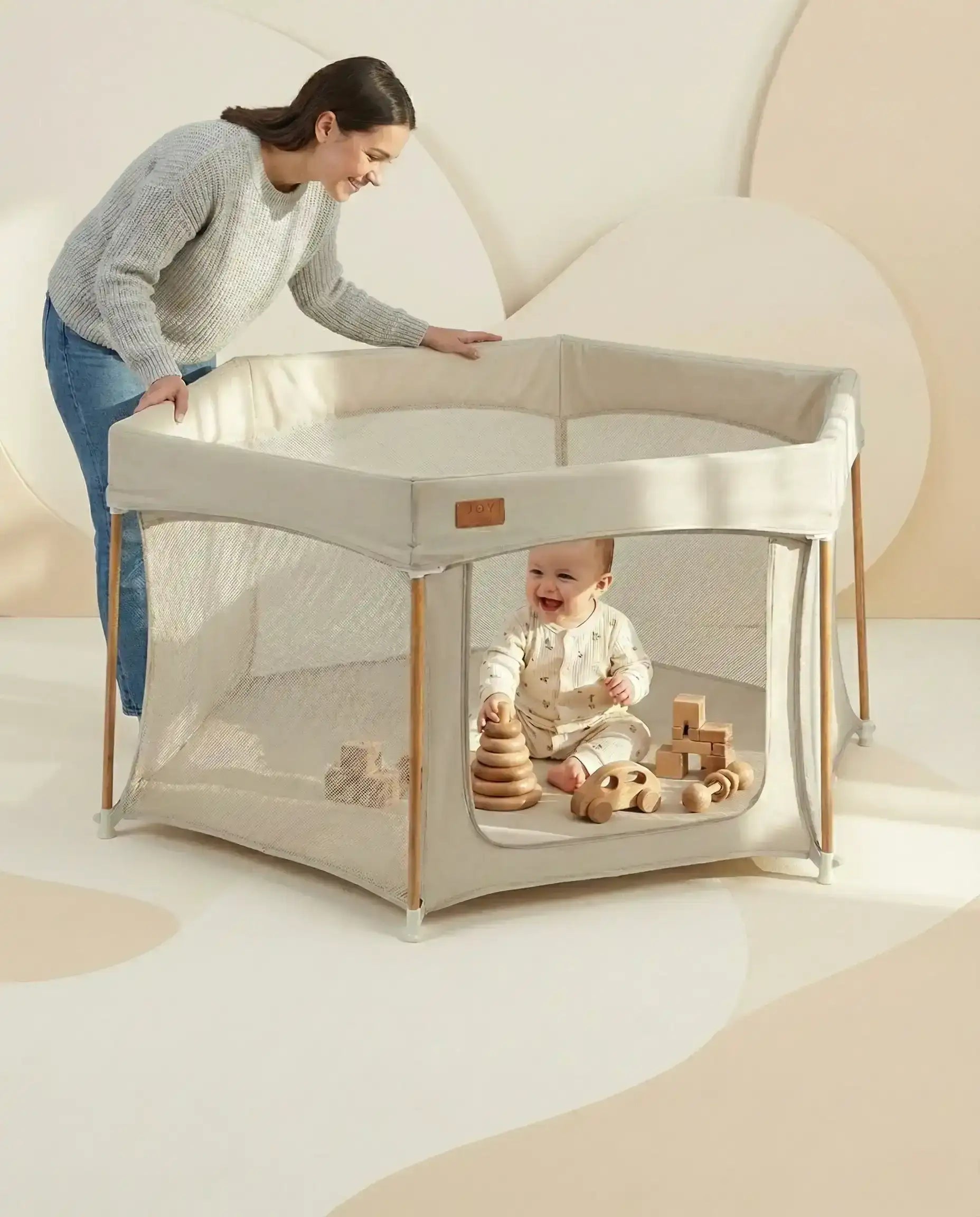 All Stars JOY Baby Playpen Natural Edition by Venture. Baby playing with wooden toys inside a portable playpen with a woman standing nearby. 
