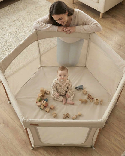 All Stars JOY Baby Playpen Natural Edition by Venture. Baby sitting in a playpen with wooden toys, watched by a woman.