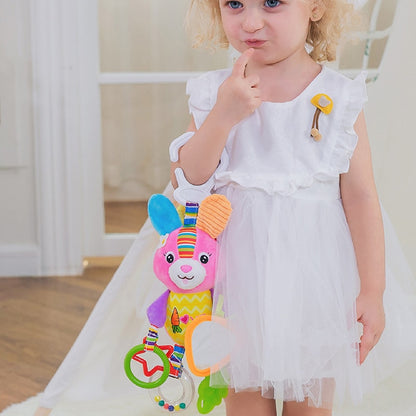 Child in a white dress holding a colorful baby toy indoors. Baby Stroller & Crib Toy Hanging Plush Sensory Animals bleu ribbon
