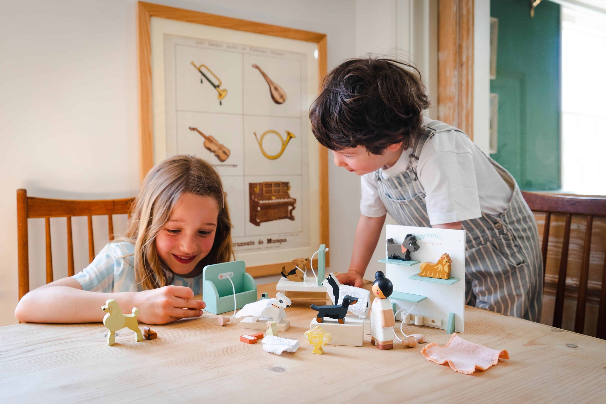Two children playing with toys at a table in a room with a decorative wall poster. Waggy Tails Dog Salon by Tender Leaf Toys - Wooden dog grooming set with 7 breeds