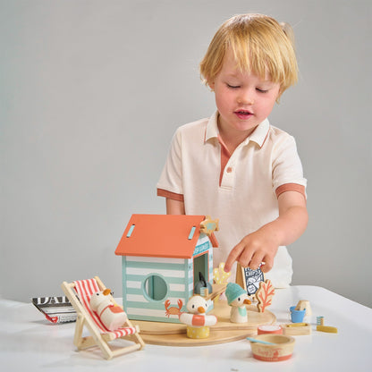 Child playing with a wooden toy house and accessories on a white surface. Sandy's Beach Hut by Tender Leaf Toys - Wooden fish and chips stall set