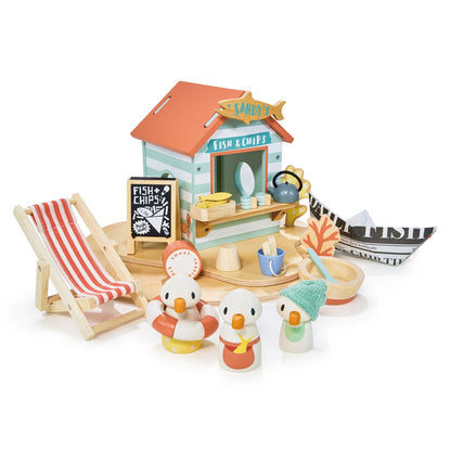 Toy set with a fish and chip shop, deck chair, and penguin figures on a white background. Sandy's Beach Hut by Tender Leaf Toys - Wooden fish and chips stall set