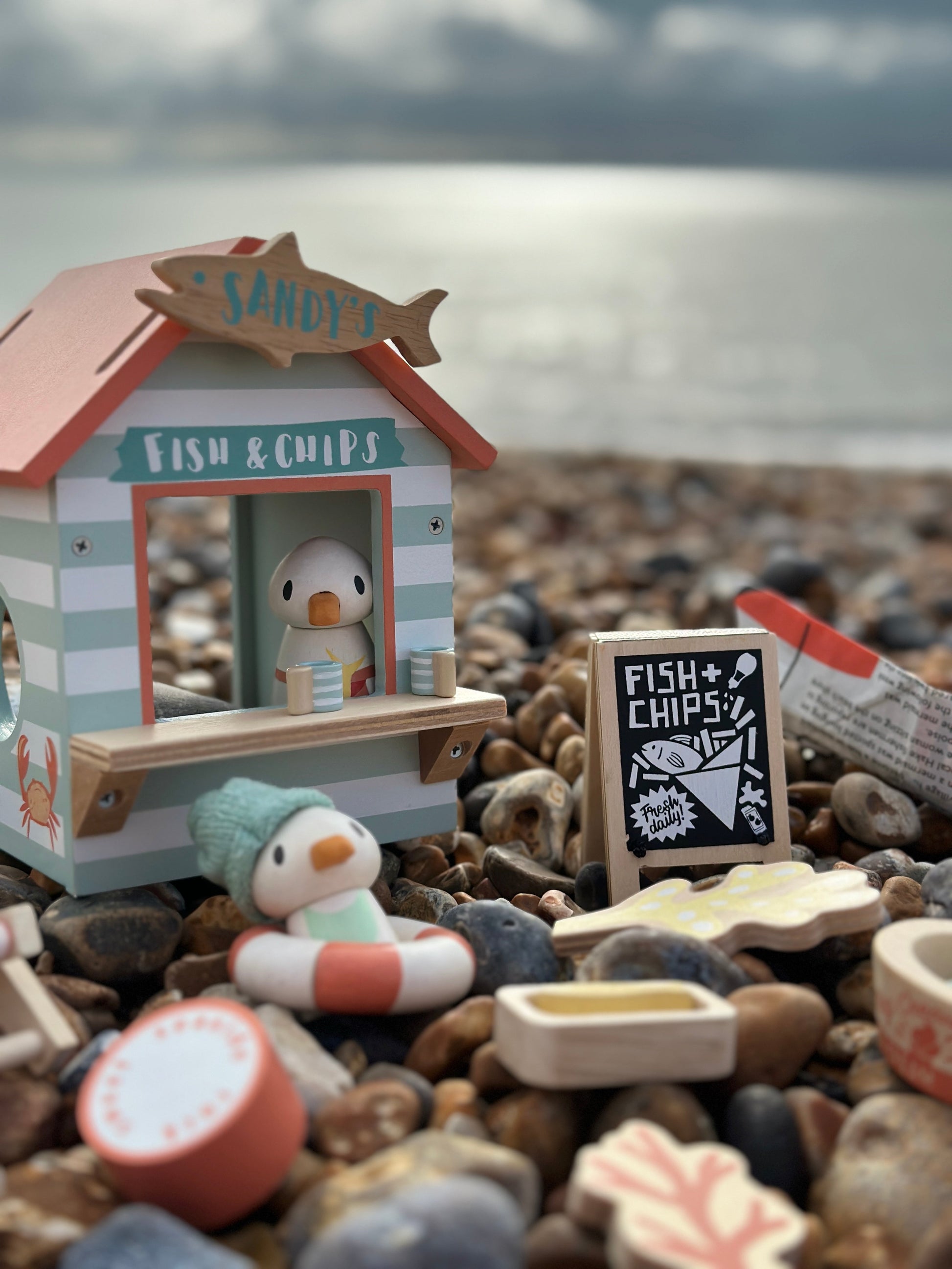 Miniature toy fish and chip shop on a pebble beach with ocean in the background. Sandy's Beach Hut by Tender Leaf Toys - Wooden fish and chips stall set