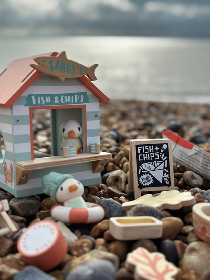 Miniature toy fish and chip shop on a pebble beach with ocean in the background. Sandy's Beach Hut by Tender Leaf Toys - Wooden fish and chips stall set