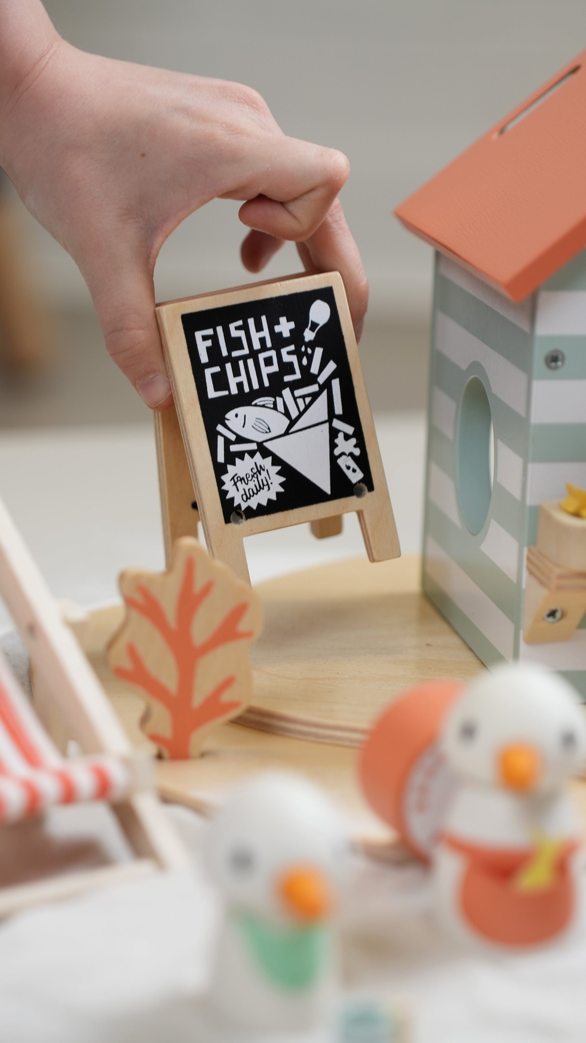 Hand holding a small wooden sign with 'Fish & Chips?' text in a playroom setting. Sandy's Beach Hut by Tender Leaf Toys - Wooden fish and chips stall set