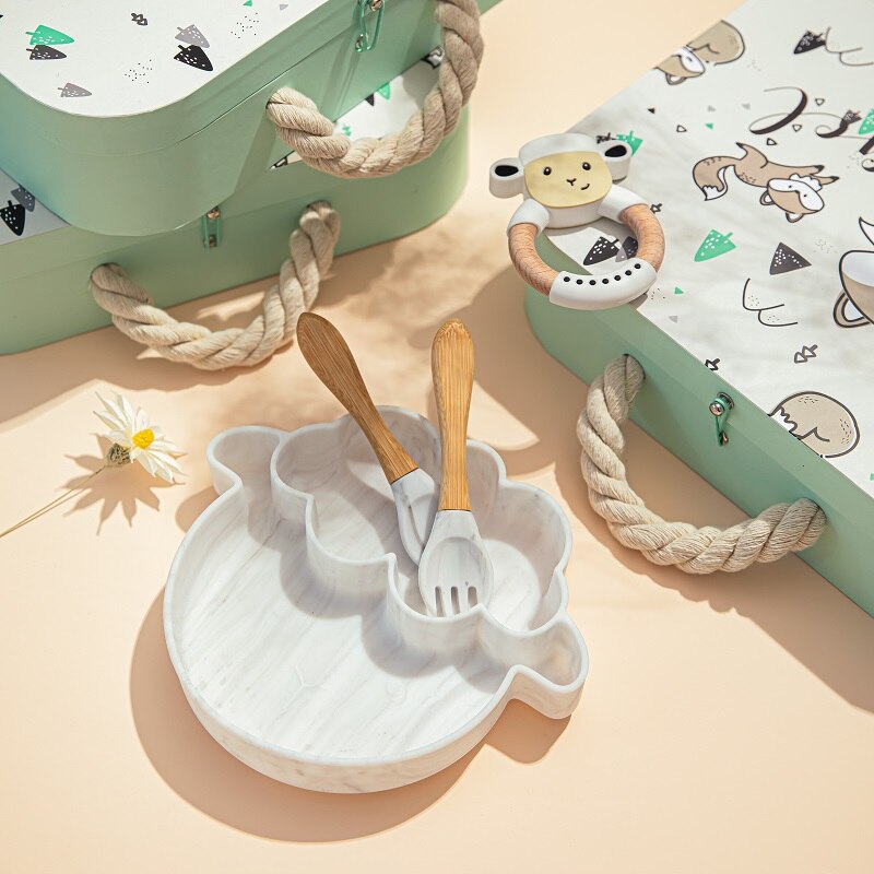 Baby utensils and toys on a light surface with a green container and patterned mat. bleu ribbon baby