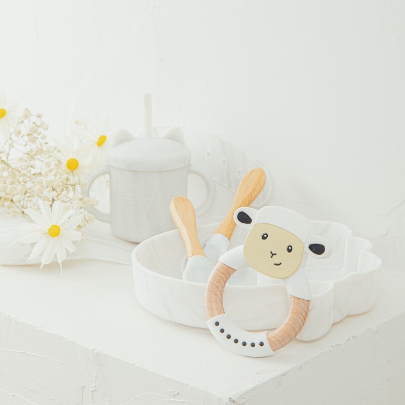 White sheep-shaped teething ring with wooden handle on a light surface with flowers in the background bleu ribbon