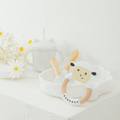 White sheep-shaped teething ring with wooden handle on a light surface with flowers in the background bleu ribbon