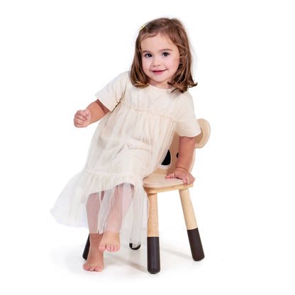 Young girl sitting on a wooden chair wearing a white dress with sheer overlay. Close-up of the hand-finished koala face back panel on the wooden nursery chair.
