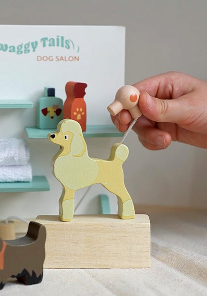 Wooden dog toy on a stand with a hand pulling a string, set against a background of 'Waggy Tails Dog Salon'. Waggy Tails Dog Salon by Tender Leaf Toys - Wooden dog grooming set with 7 breeds