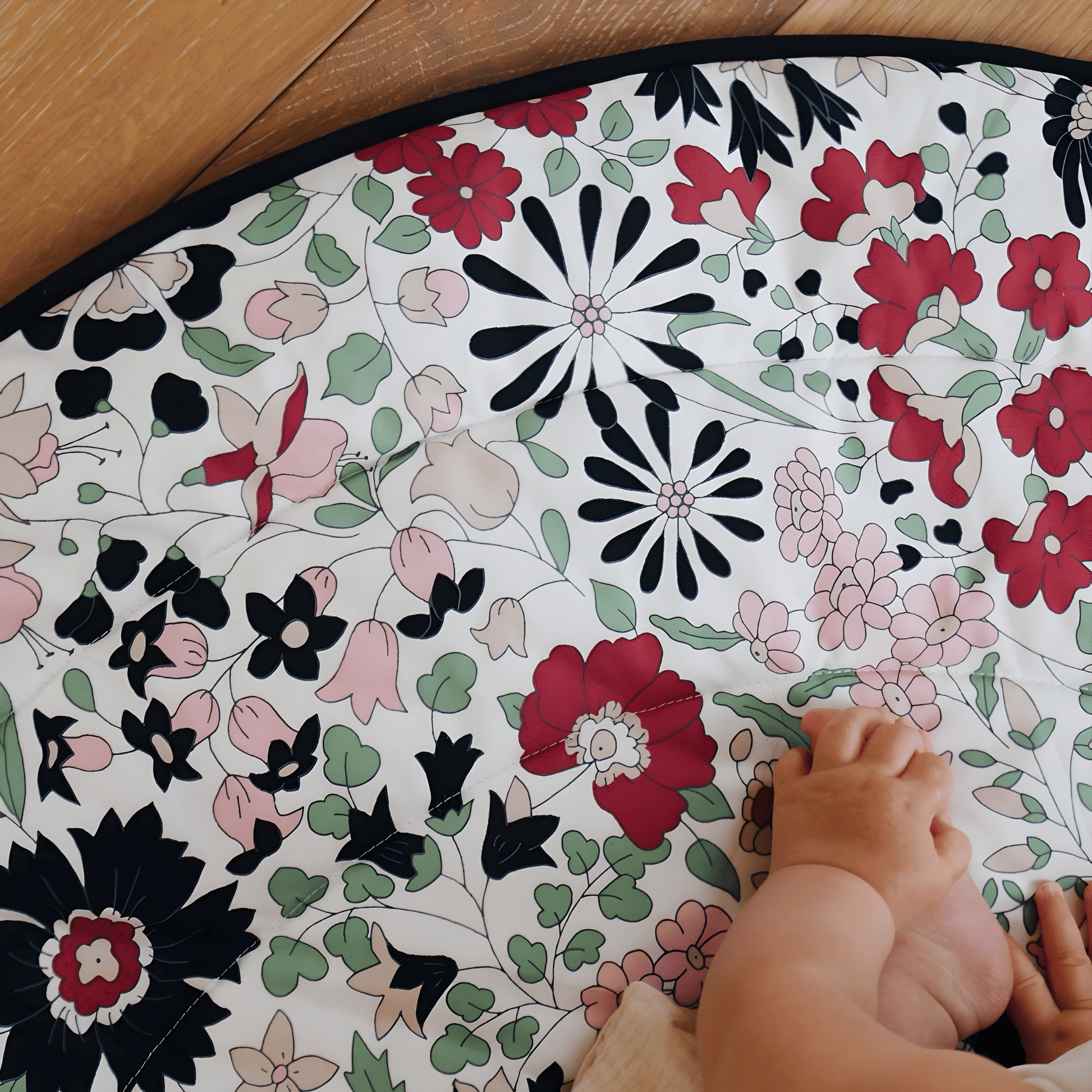 Close up of a baby's legs resting on the vibrant floral pattern of the Etta Loves x Liberty reversible playmat on a wooden floor.
