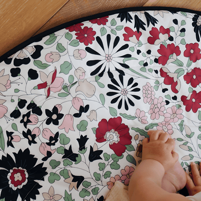 Close up of a baby's legs resting on the vibrant floral pattern of the Etta Loves x Liberty reversible playmat on a wooden floor.
