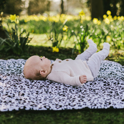 A baby lying comfortably on the back, looking around while resting on the high-contrast Etta Loves x Liberty outdoor sensory playmat in a sunny park with daffodils.