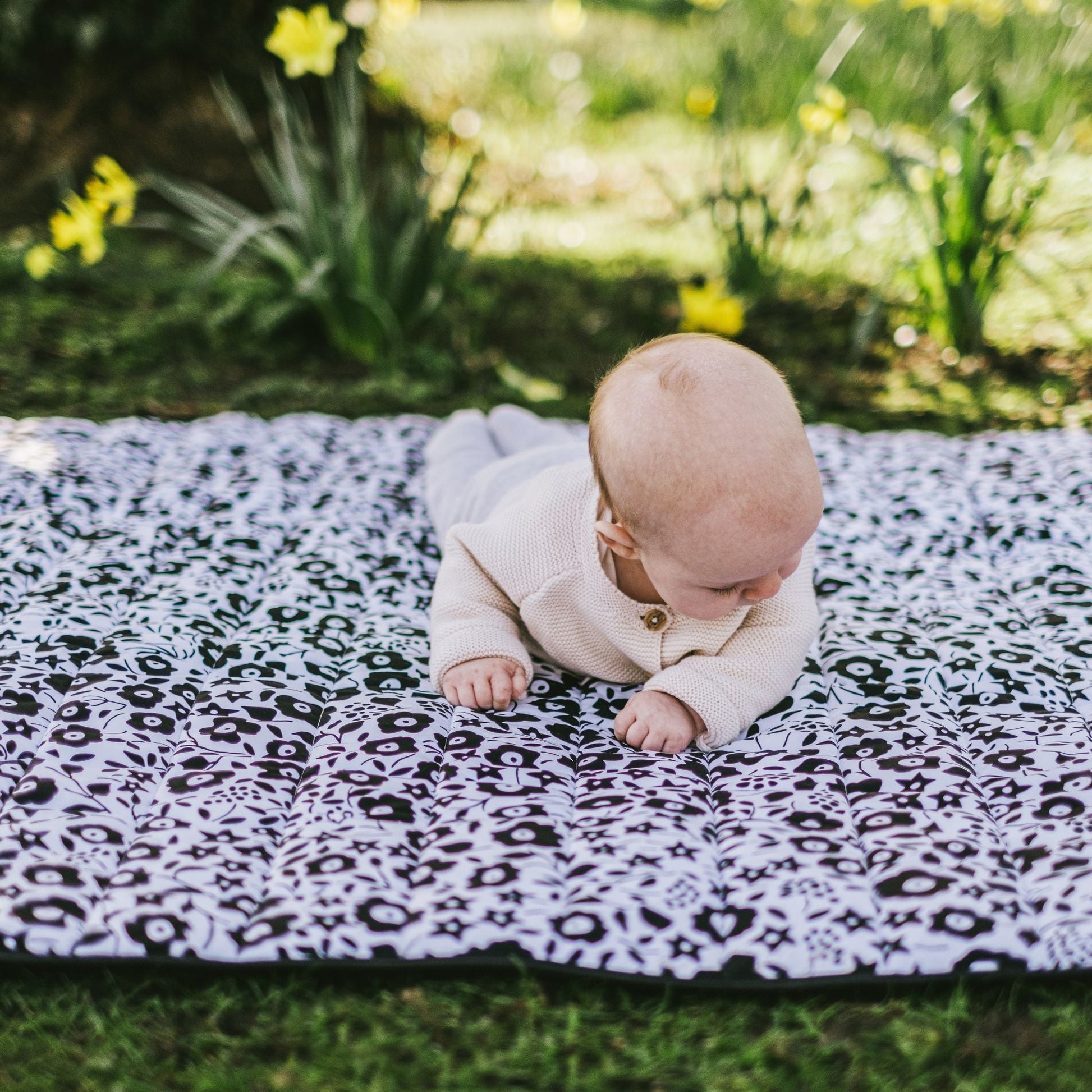 A baby practicing tummy time and developing neck strength on the visually stimulating Etta Loves x Liberty outdoor playmat outdoors among spring flowers.