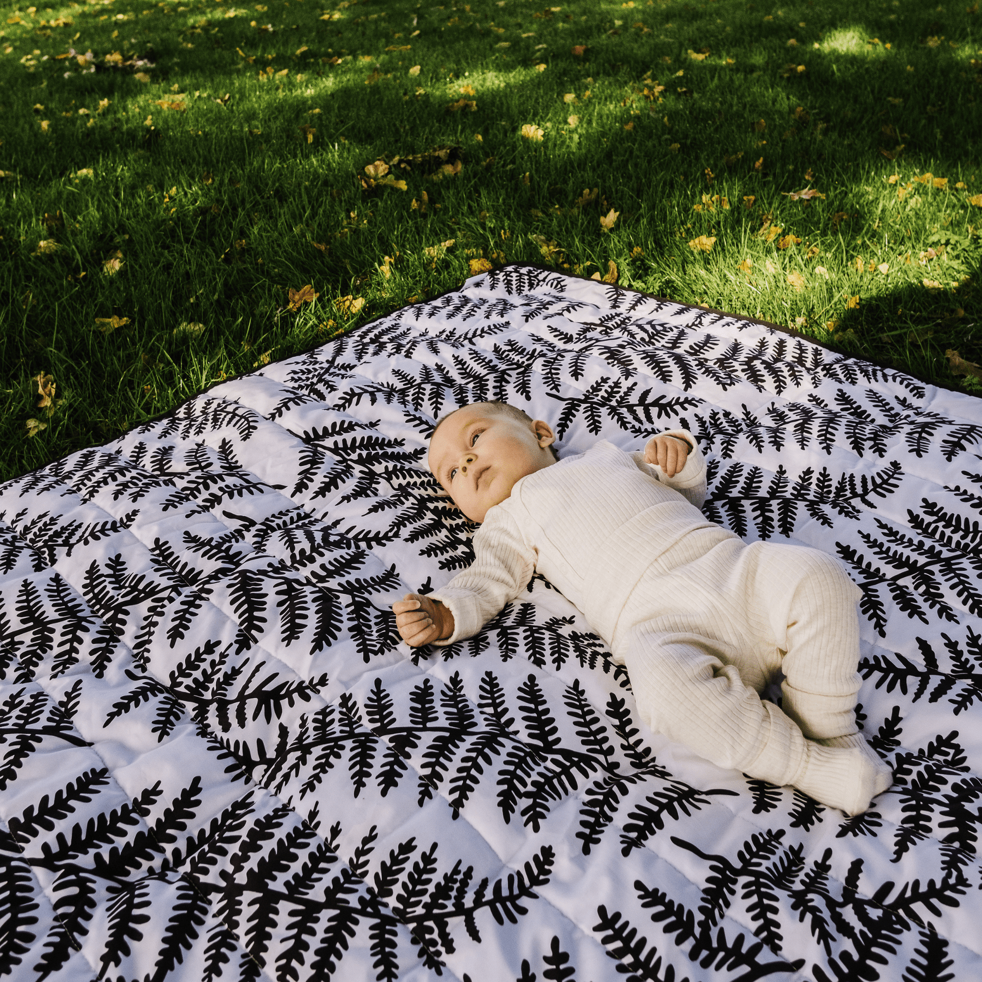 A baby engaging with the black and white fern pattern of the Etta Loves outdoor sensory playmat on green grass.