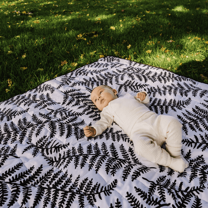 A baby engaging with the black and white fern pattern of the Etta Loves outdoor sensory playmat on green grass.