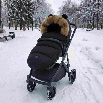 Black Nordic winter stroller cocoon attached to a pram in a snowy park. Windproof baby footmuff with cute bear ears and faux fur hood for cold weather walks.