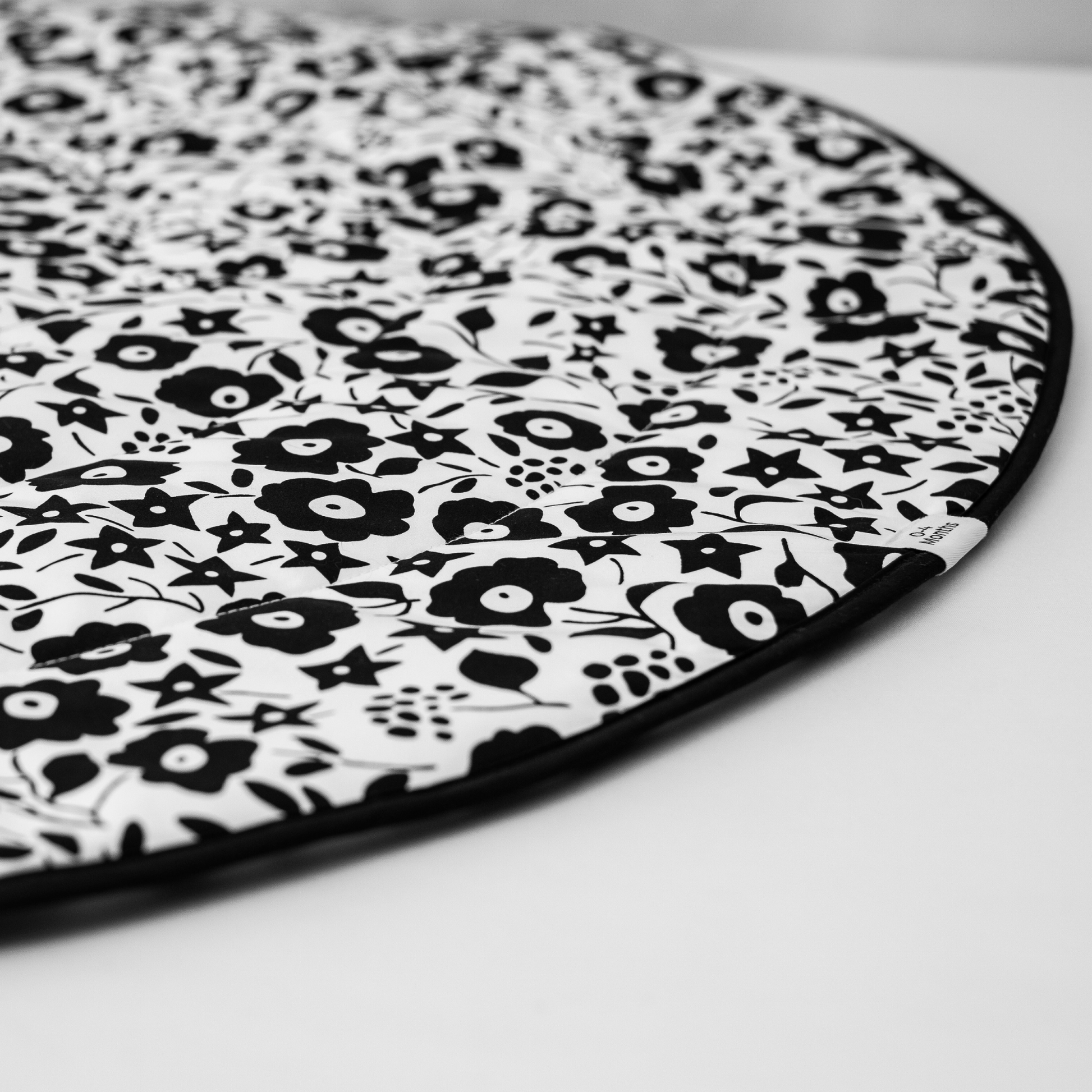 Close-up of the softly padded edge of the Etta Loves round playmat featuring the 0-4 Months developmental stage tag on the black and white Liberty print.