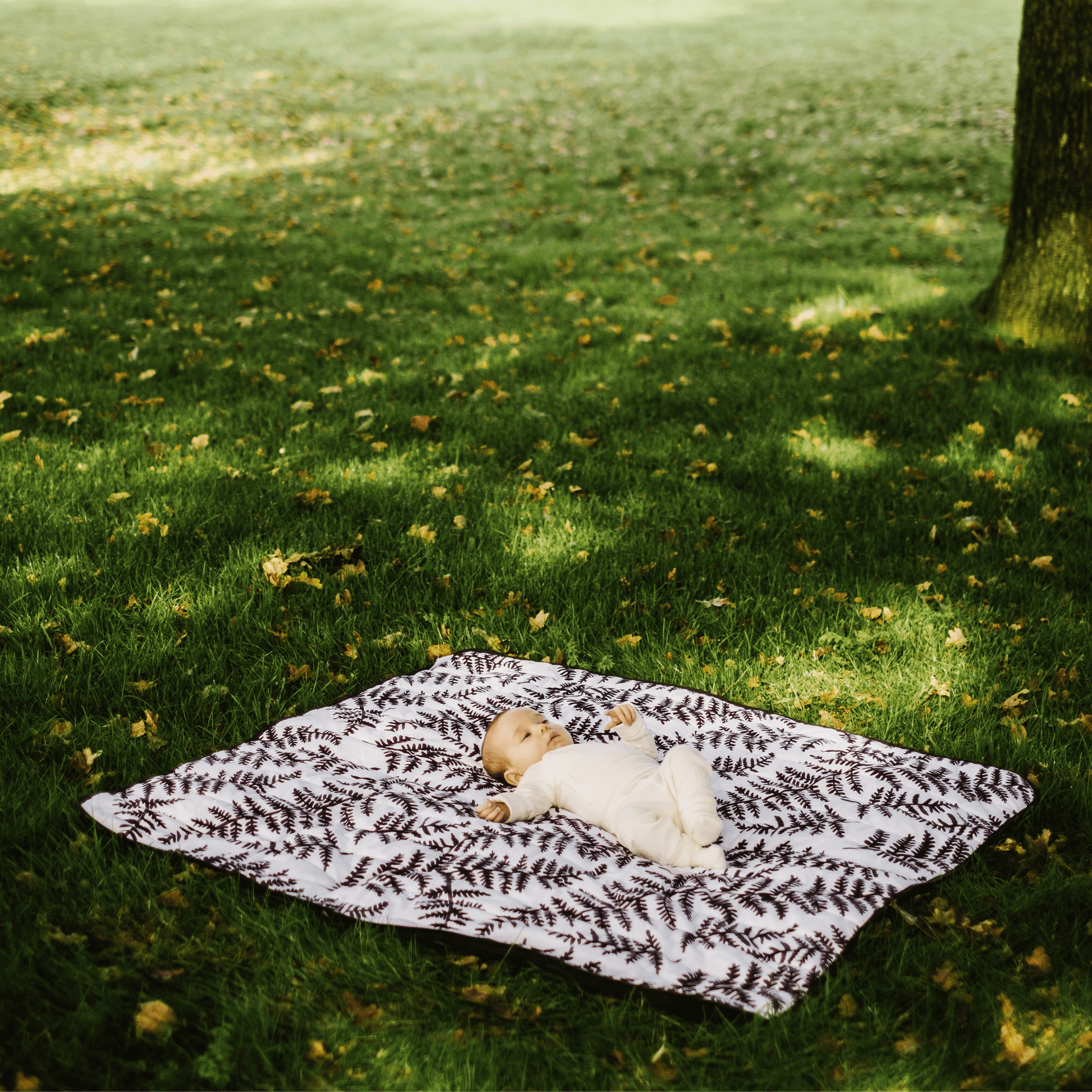 Wide shot of an infant resting comfortably on the square, softly padded Etta Loves fern print outdoor playmat under the shade of a tree.