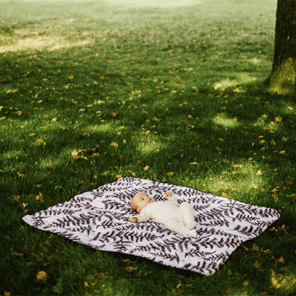 Wide shot of an infant resting comfortably on the square, softly padded Etta Loves fern print outdoor playmat under the shade of a tree.