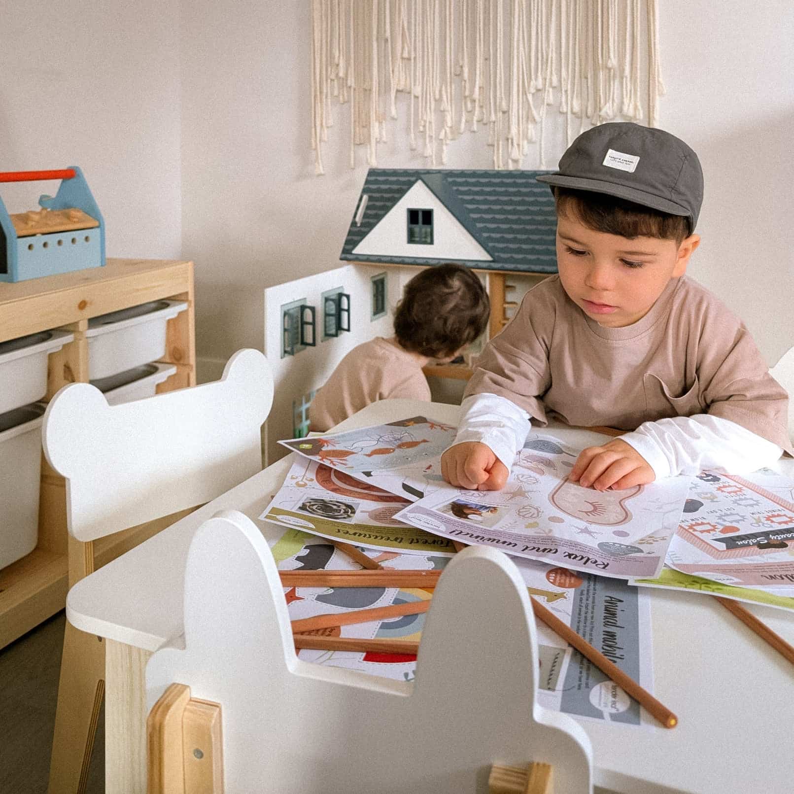 Two children playing with a toy house and magazine in a room with white walls and wooden furniture. Classic wooden nursery table for four children group play