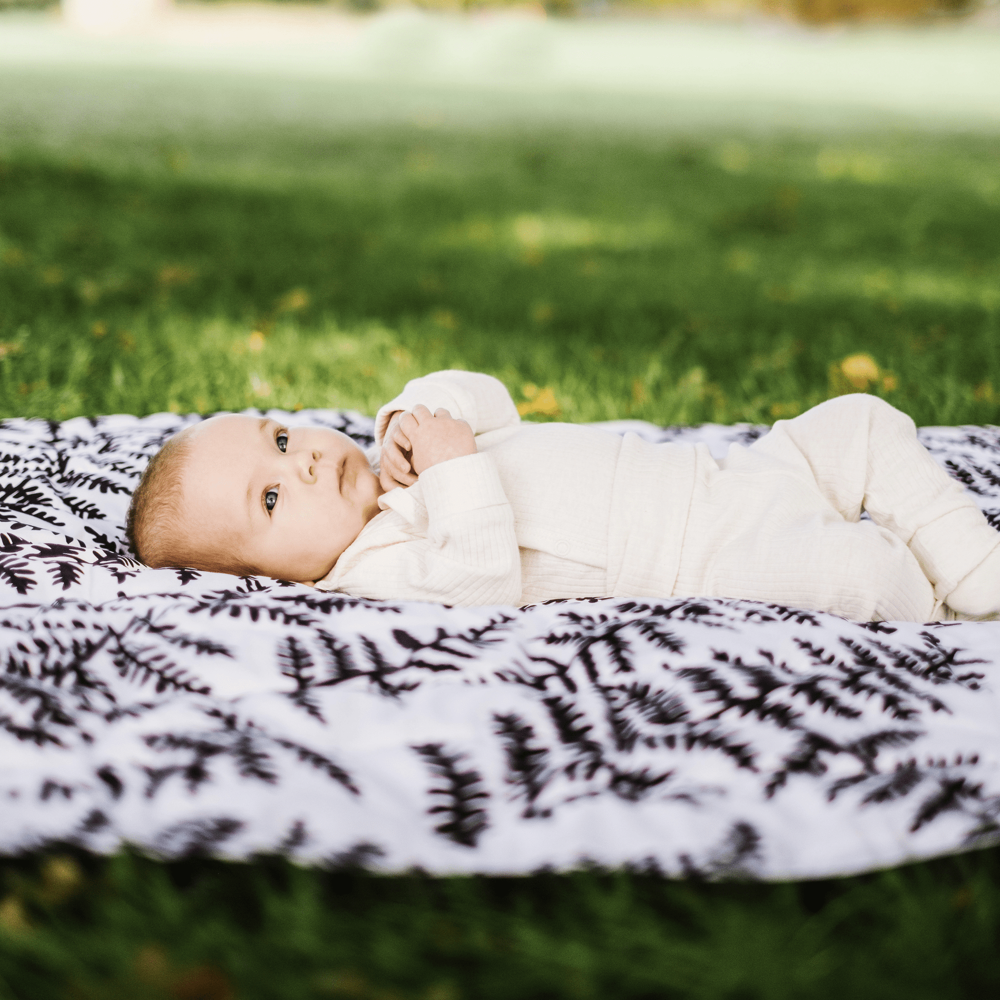 A calm newborn baby lying on the high-contrast Etta Loves fern print sensory playmat in a grassy park setting.