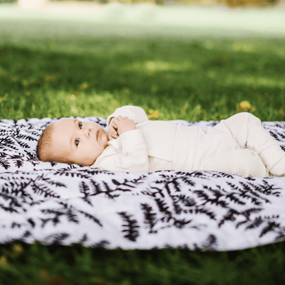 A calm newborn baby lying on the high-contrast Etta Loves fern print sensory playmat in a grassy park setting.