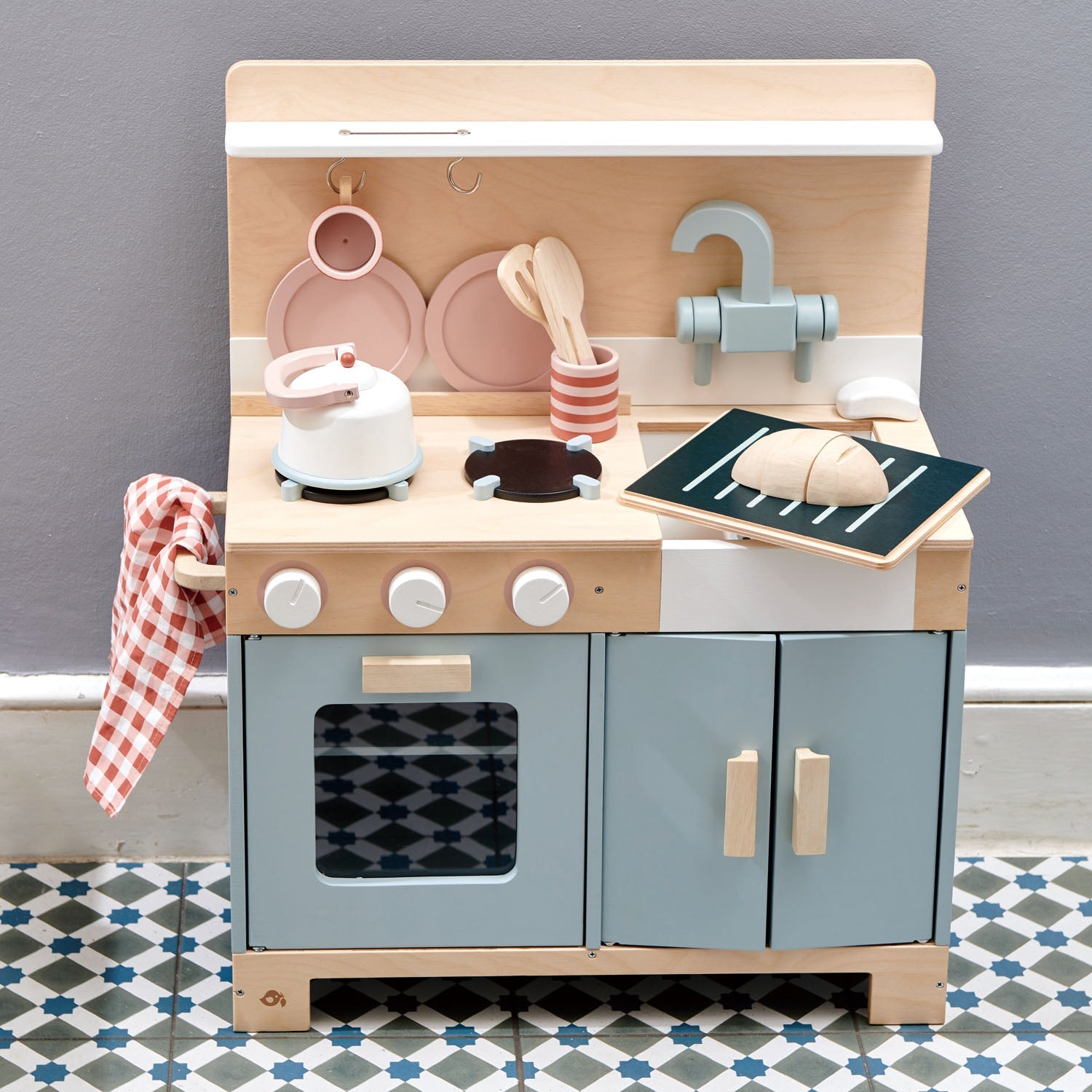Wooden toy kitchen set with various accessories on a gray wall background the play kitchen featuring an opening oven door with shelf and a spacious storage cupboard under the sink for toy organization.