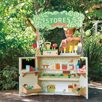 Child playing at a toy store set outdoors with greenery in the background Wooden play shop interior with shelves, strawberry tote bag, and open doors for grocery play.